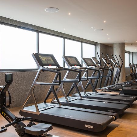 Отель Grand Hyatt Барселона Экстерьер фото The photo shows a modern gym space featuring several pieces of exercise equipment. In the foreground, there are treadmills, while in the background, there are elliptical machines lined up against large windows that provide natural light. The flooring